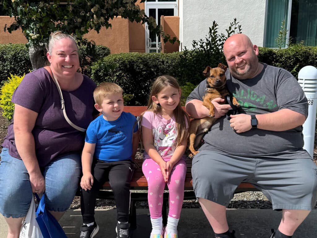 A family of four sits on a bench outside. A woman and man hold a small dog, flanked by two children. The girl wears a pink outfit, and the boy is in a blue shirt. They are smiling, surrounded by greenery, highlighting the joy of adoption.