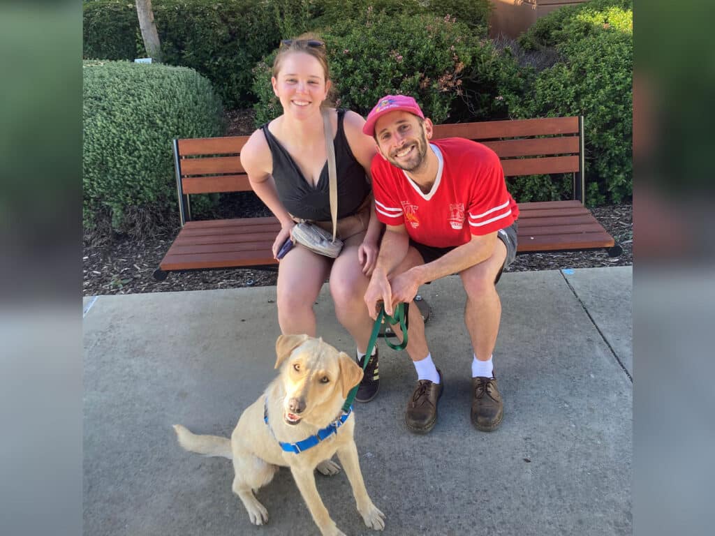 A woman and a man sitting on a bench with a light-colored dog wearing a blue collar. The woman is wearing a black outfit and the man is in a red shirt. The setting appears to be an outdoor park area, reflecting a positive moment related to adoption.