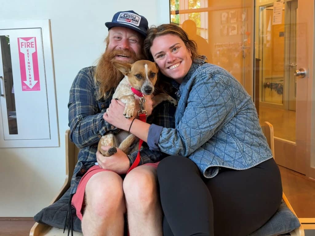 A man with a long beard and a cap sits on a chair, holding a small dog with a red collar. A woman in a blue jacket sits next to him, embracing him and the dog. They are in a cozy indoor space with bright lighting, symbolizing adoption.