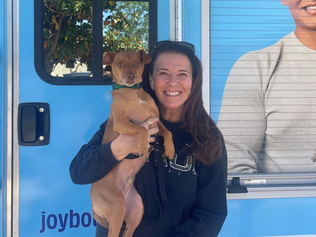 A woman in a black sweatshirt holds a small dog in front of a vehicle featuring a blue design. Both the woman and the dog appear content. The scene represents the theme of adoption, highlighting the joy of welcoming a pet into a home.