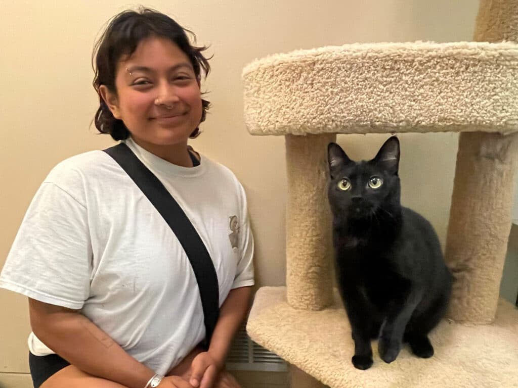 A young woman with short hair, wearing a white t-shirt and a black crossbody bag, smiles next to a black cat sitting on a beige cat tree. Both are looking toward the camera in a setting that suggests an adoption environment.