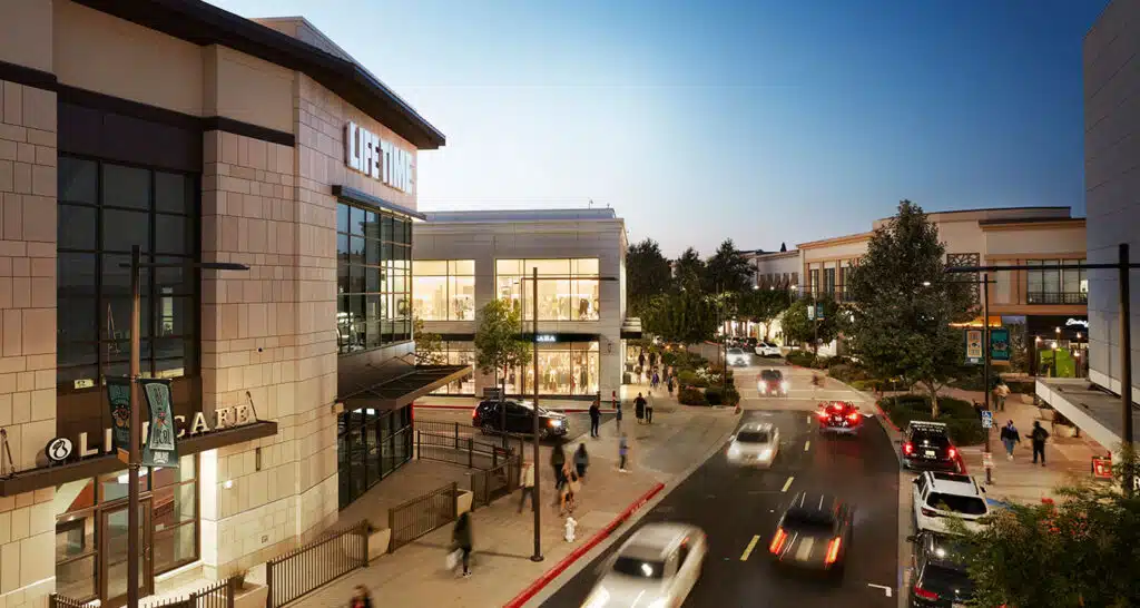 An evening view of a shopping area featuring a Lifetime gym on the left, surrounded by modern architecture, trees, and a bustling street with cars and pedestrians. The sky shows dusk lighting, creating a lively urban atmosphere.
