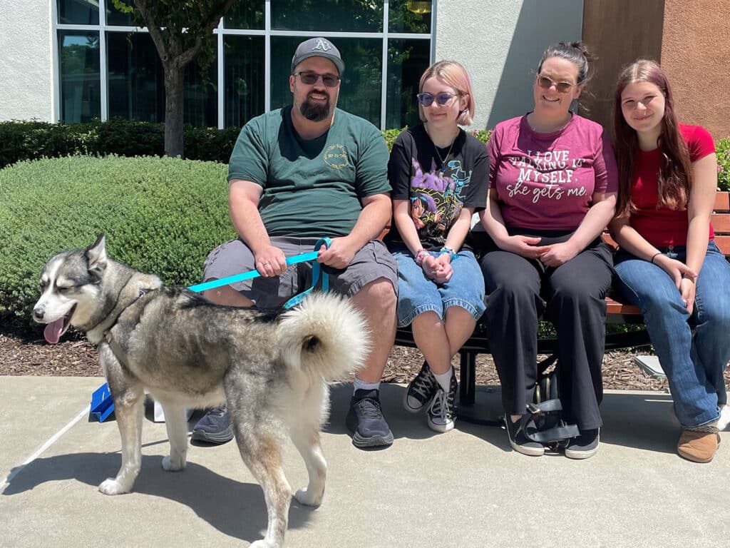 A man and three individuals sitting on a bench outside a building, smiling. A Siberian Husky on a leash stands next to them. Trees are visible in the background. This image relates to Adoption activities during the week of June 2.