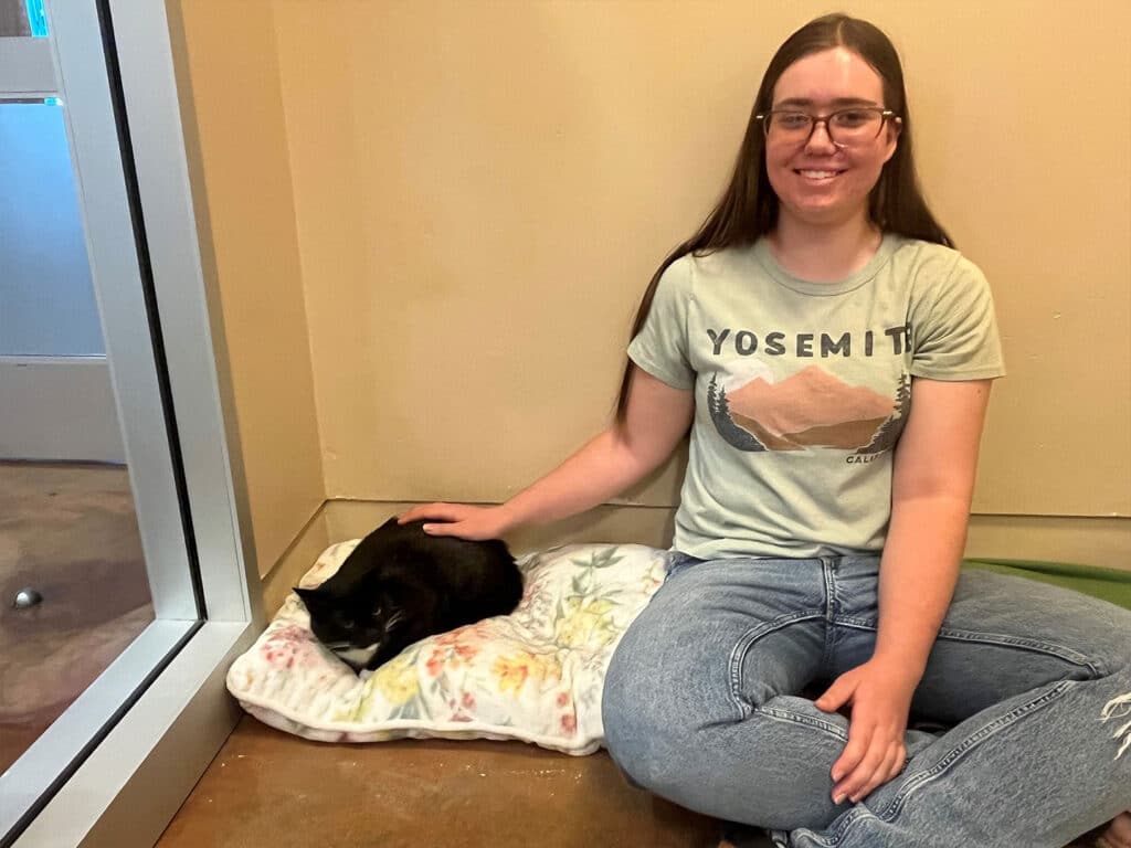 A young woman wearing glasses and a Yosemite-themed t-shirt sits on the floor beside a black cat resting on a soft blanket. She gently strokes the cat, which appears relaxed in the cozy indoor setting. The image illustrates a moment during an adoption event.