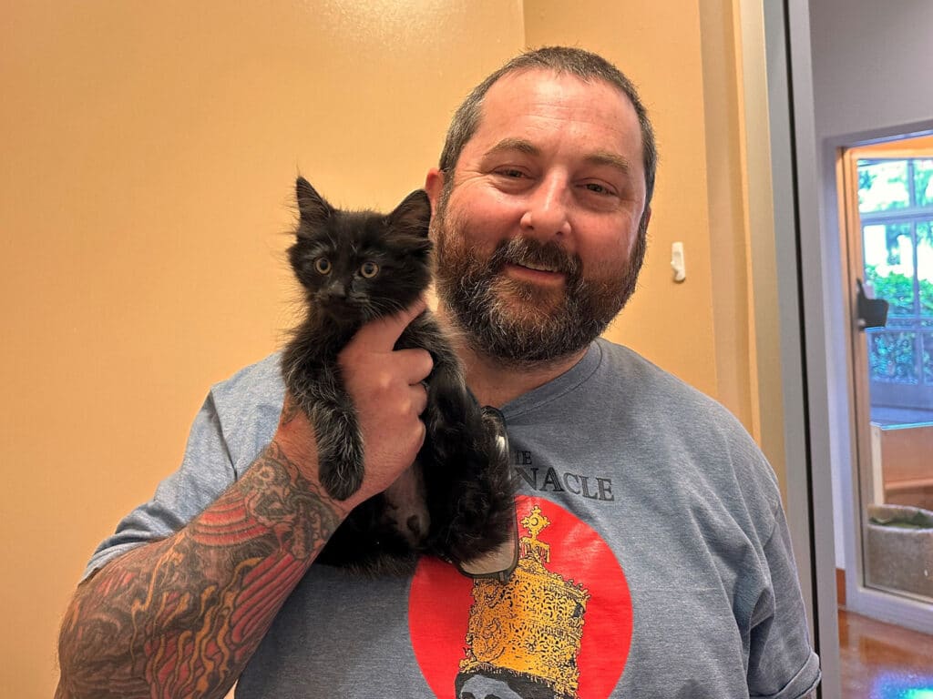 A man with a beard and tattoos holds a small black kitten close to his chest, smiling. They are indoors, with a light-colored wall and window visible in the background. This image highlights a moment of joy related to pet adoption.
