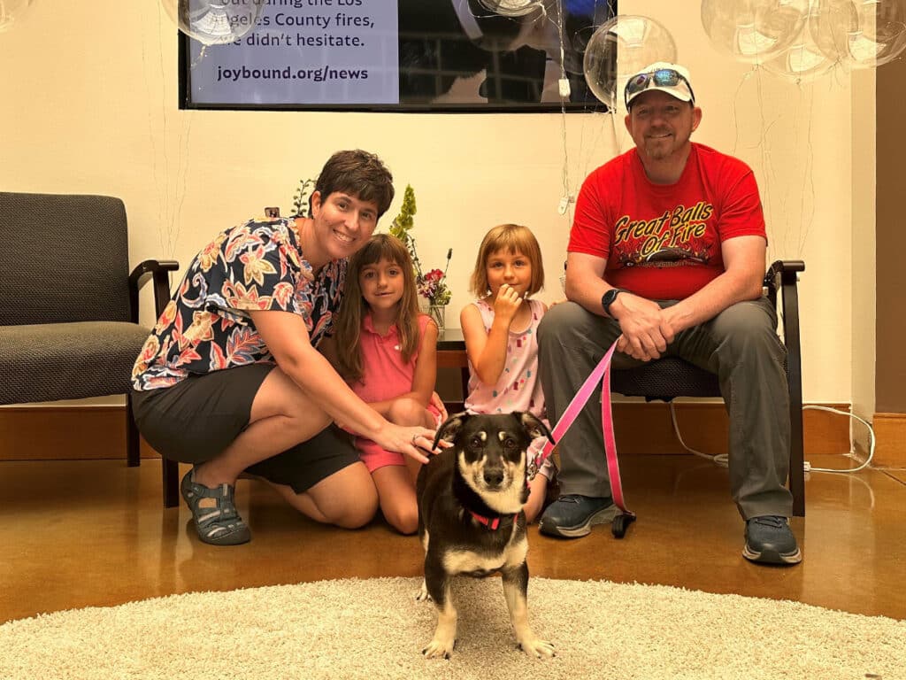 A family of four is seated in a pet adoption center. A woman and two young girls sit on the floor next to a small dog on a leash. A man is sitting beside them, smiling. The background features a screen displaying adoption information.