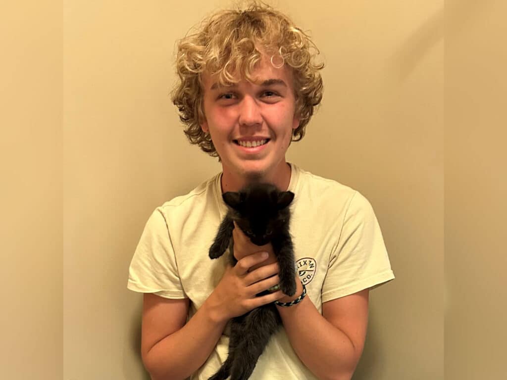A young man with curly hair smiles while holding a black kitten in front of a plain wall. The image highlights a positive moment related to pet adoption.