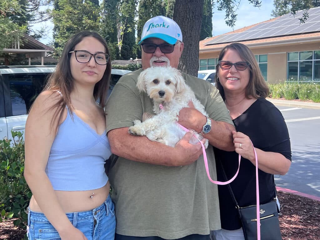 A man wearing a blue hat holds a small dog while standing between two women outside. The woman on the left wears a light blue top, and the woman on the right is in a black shirt. They are smiling in a parking area, indicating an adoption event.