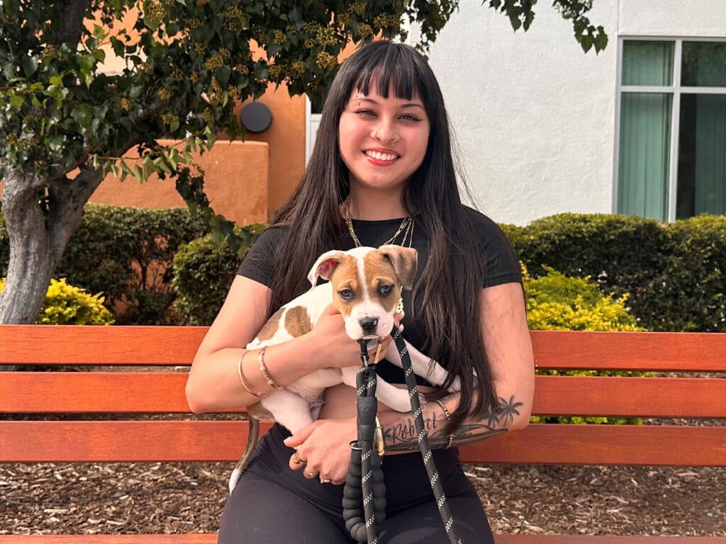 A young woman with long black hair sits on a wooden bench, holding a small puppy with a white and brown coat. Green bushes and a building are in the background, depicting a pleasant outdoor setting, highlighting the theme of Adoption.