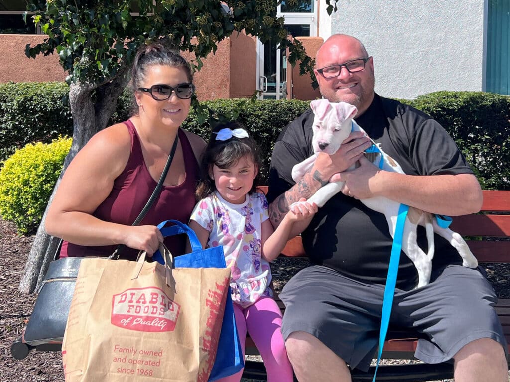 A family sits on a bench outside, smiling. The mother holds a bag, while the father holds a puppy on a leash. A young girl, wearing a shirt with colorful designs, sits between them, gently holding the puppy. The background features greenery and a building.