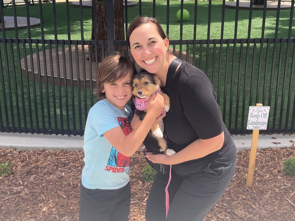 A woman and a child stand outside in a fenced area, both smiling. The child holds a small puppy, wearing a pink bandana, while the woman, dressed in black, supports the puppy. Green grass is visible in the background, related to adoption.