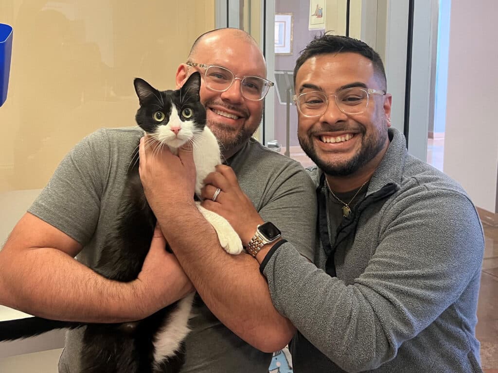 Two men smiling while holding a black and white cat in an adoption center. The men are wearing gray sweaters and glasses, standing in front of a window with light coming through, celebrating the cat's adoption.