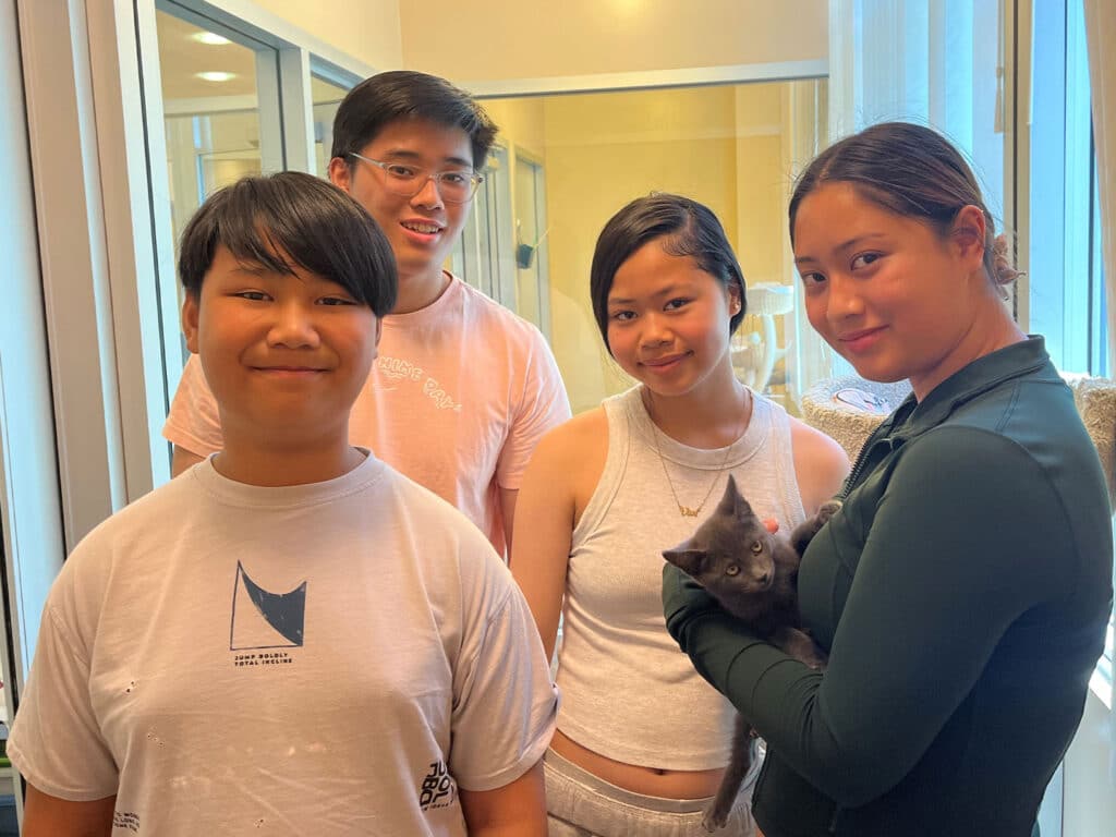 Four young people are gathered in a bright room, smiling at the camera. One holds a gray kitten. The scene captures a moment of joy and connection related to adoption, emphasizing the bond formed between the youth and their new pet.