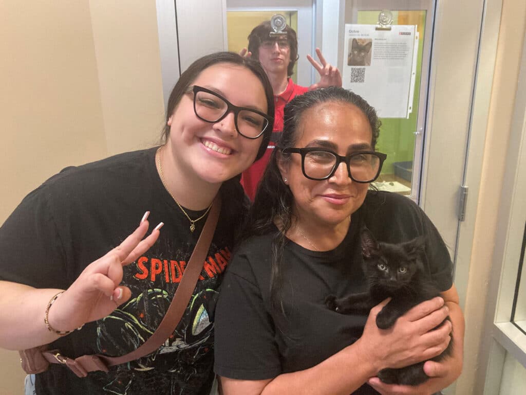 Two women pose for a photo in an animal adoption center. One woman holds a small black kitten, while the other makes a peace sign. A young man in the background gives a playful expression. The setting appears bright and welcoming.
