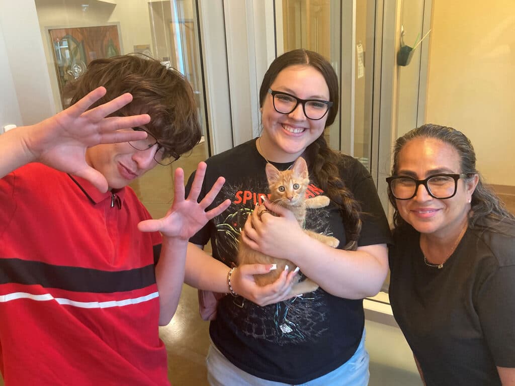 Two people smiling in an animal shelter with a small orange kitten. One person is wearing glasses and holding the kitten, while the other, in a red shirt, playfully poses beside them. A woman with glasses stands to the right, smiling. This scene supports the theme of adoption.
