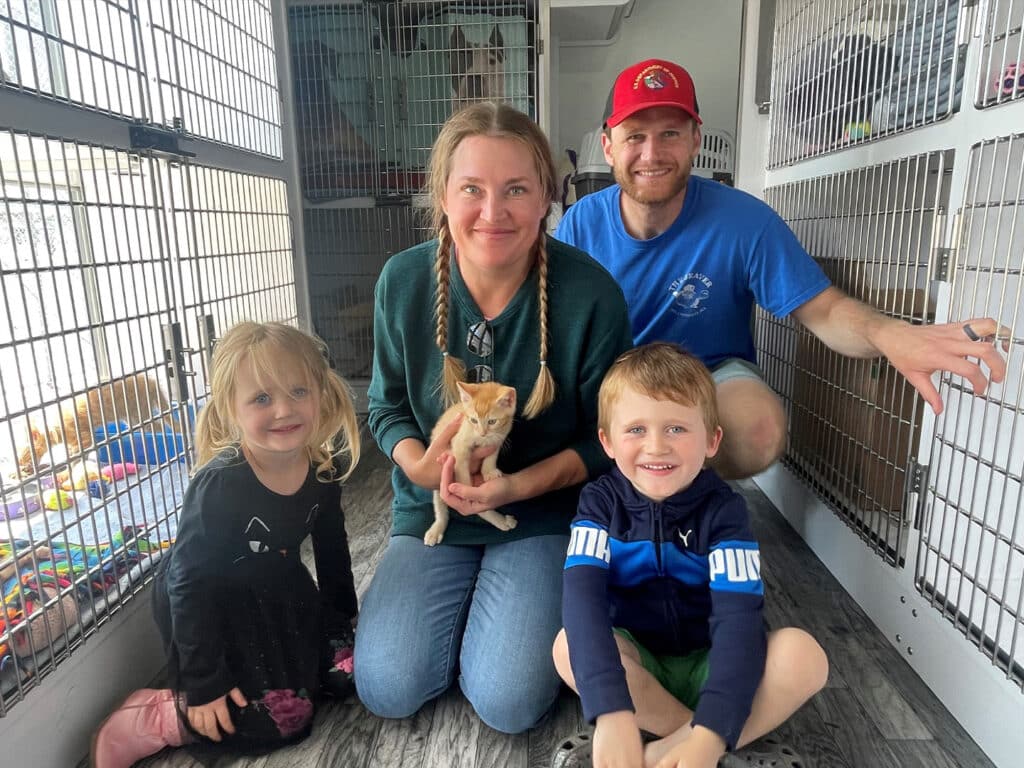 A family with two children and one adult is posed in an animal shelter, each smiling and kneeling on the floor. The adult holds an orange kitten, and behind them are animal cages. The scene reflects a moment of joy related to pet adoption.