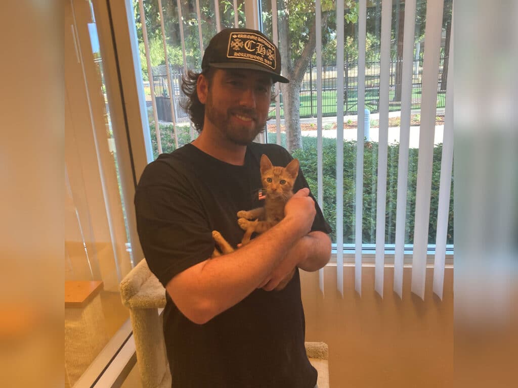A man wearing a black shirt and cap holds a small orange kitten in his arms inside a bright room with large windows and green outdoor space visible. This image highlights the joy of pet adoption.