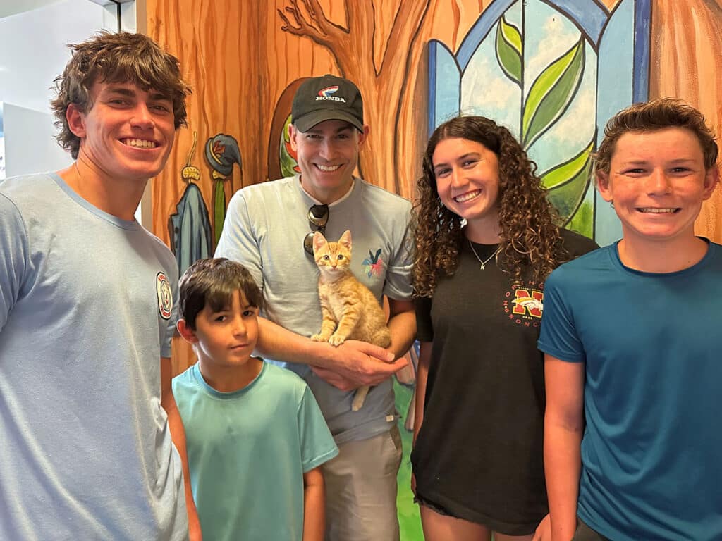 A group of five people stands in front of a colorful mural. A man holds an orange kitten while four individuals smile around him, including two young men, a young girl, and a young woman. The setting suggests a supportive adoption environment.