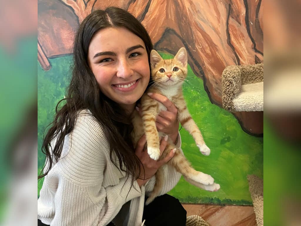A woman holding an orange tabby kitten in front of a colorful mural depicting green grass and trees. Both the woman and kitten are looking at the camera, highlighting a moment of companionship related to pet adoption.