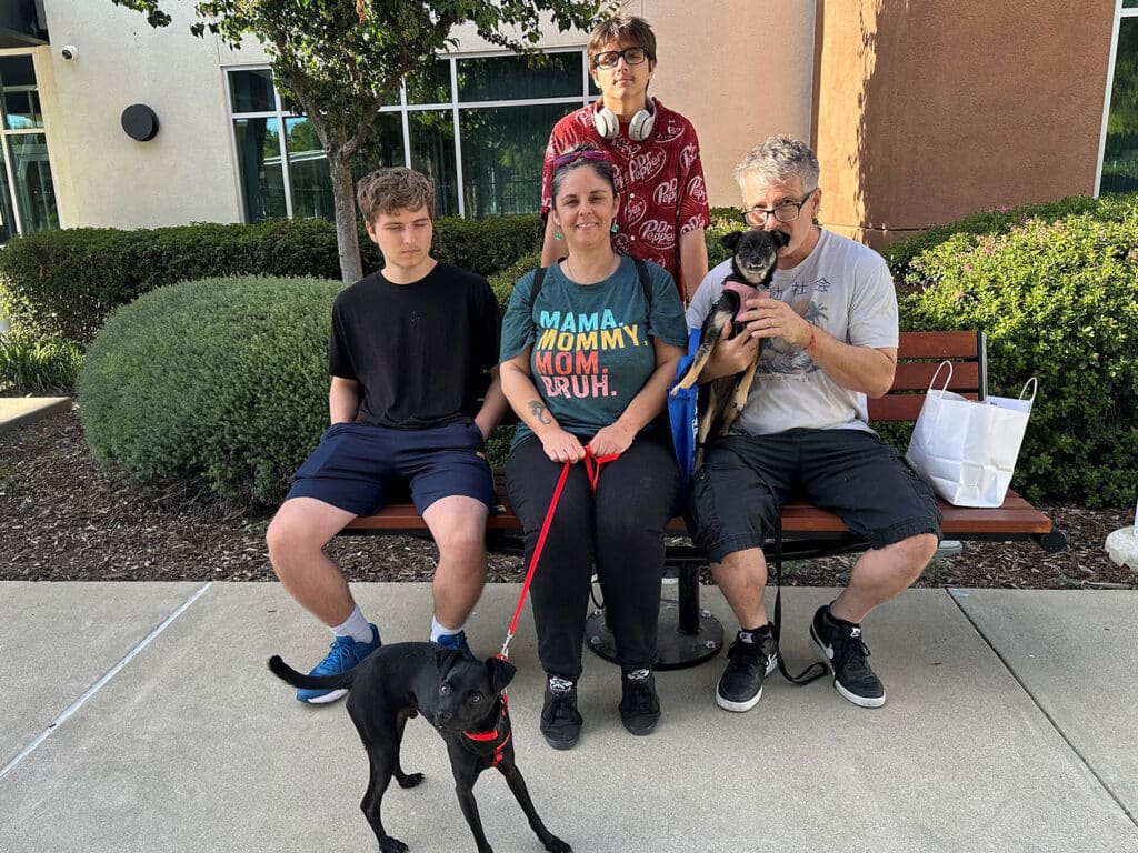 A family sits on a bench outside, consisting of three adults and two teenagers. One woman is holding a small dog, while another dog is standing in front on a leash. The group appears cheerful and is surrounded by greenery.