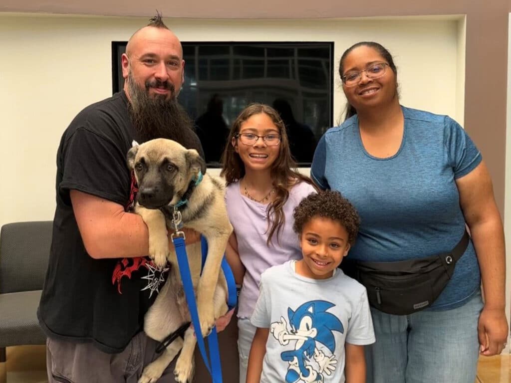 A family poses together in a room, smiling with a large dog. The father holds the dog in his arms, while a girl with glasses stands next to him, a boy in a blue shirt smiles in front, and a woman stands on the right. This image highlights an adoption moment.
