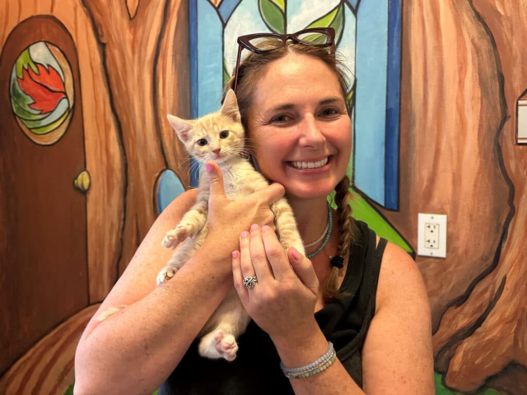 A woman with long brown hair, wearing a black top, smiles while holding an orange kitten against a colorful mural of a tree. The setting appears to be a playful space, highlighting the theme of adoption.