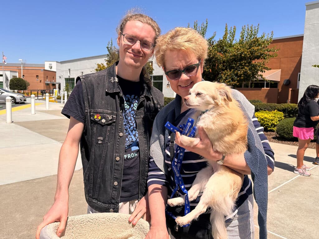 A young man and a woman holding a small dog outside a shelter. The man, wearing a black denim vest, stands beside the woman who has short blonde hair and glasses. The dog is on a blue leash, and they appear to be participating in an adoption event.