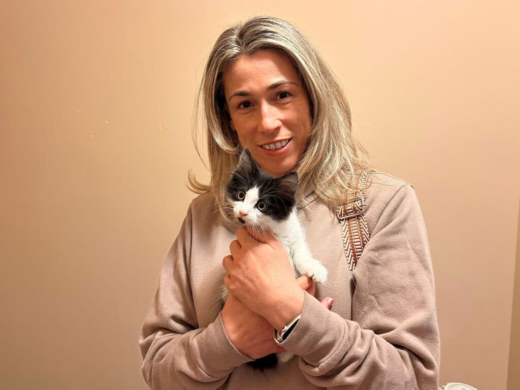 A woman with long, light-colored hair holds a small black and white kitten in her arms. The setting features a simple interior wall. This image relates to adoption, highlighting the bond between the woman and the kitten.