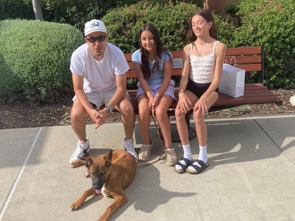 A man and two girls sit on a bench in a park, with a brown dog lying on the ground in front of them. The man wears sunglasses and a white cap, while one girl has long hair and the other has a ponytail. They are gathered in a friendly setting, highlighting the theme of Adoption.