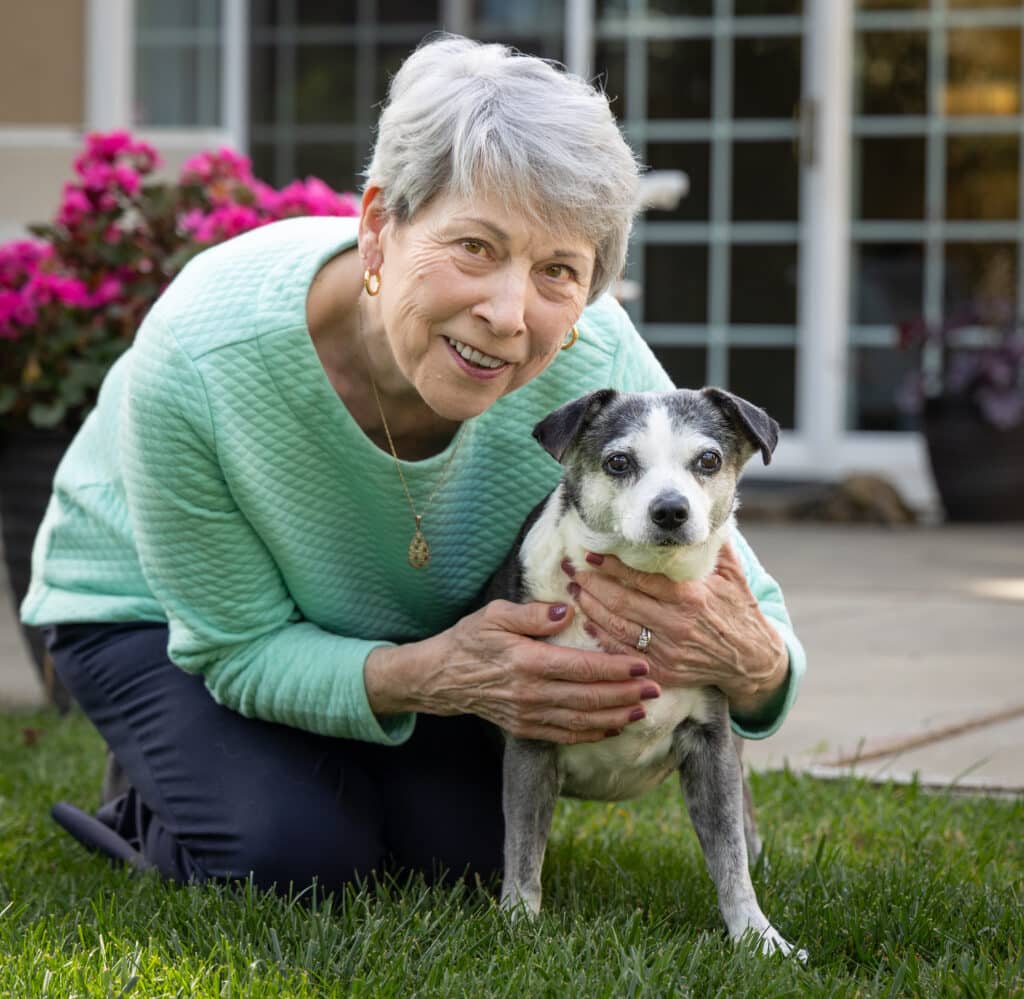 An older woman with short gray hair, wearing a light blue sweater, crouches on a grassy area holding a small black and white dog. Pink flowers are visible in the background, and a house with large windows is behind them.