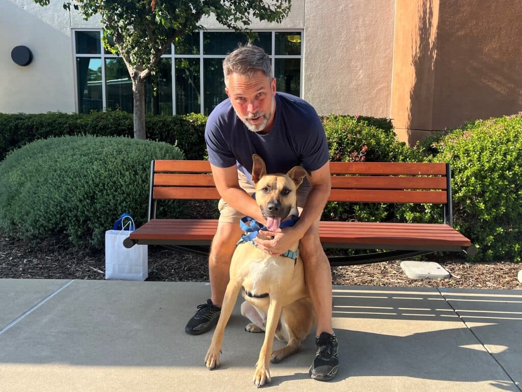 A man sitting on a wooden bench outdoors with a dog. The man, wearing a dark shirt and shorts, is leaning forward with a playful expression, while the dog, a tan-colored mixed breed, sits on the ground next to him with a blue collar.