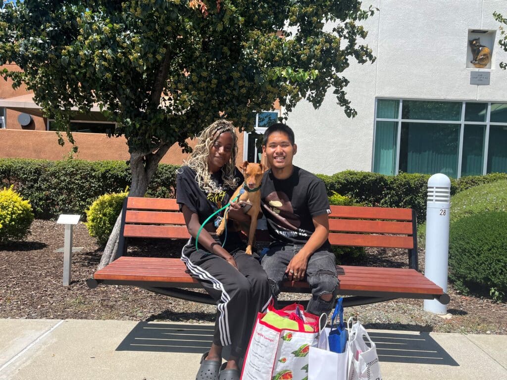 A woman and a man sitting on a bench outside, holding a small dog. The woman has long braided hair and wears a black shirt with stripes. The man is wearing a black shirt and shorts. They have bags beside them, and there are green bushes in the background.
