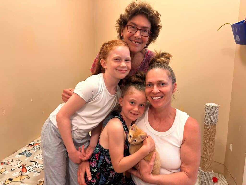 A family of four, including two girls, is gathered in a room with a beige wall. One girl holds a small orange kitten, while the other leans against her mother. The mother smiles at the camera, and the father stands behind the girls.