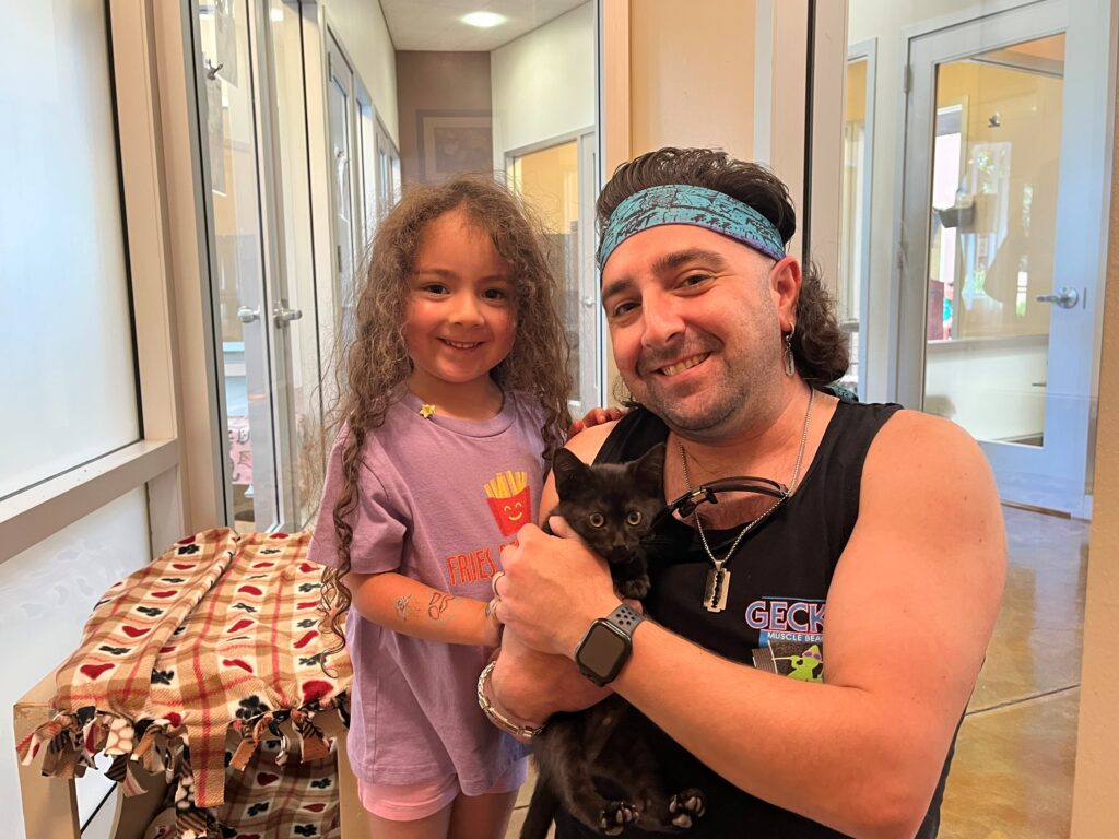 A man holding a black kitten in one arm smiles alongside a young girl with curly hair who stands beside him, also smiling. The setting is indoors, featuring large windows and light-colored walls. A patterned blanket is on a nearby table.
