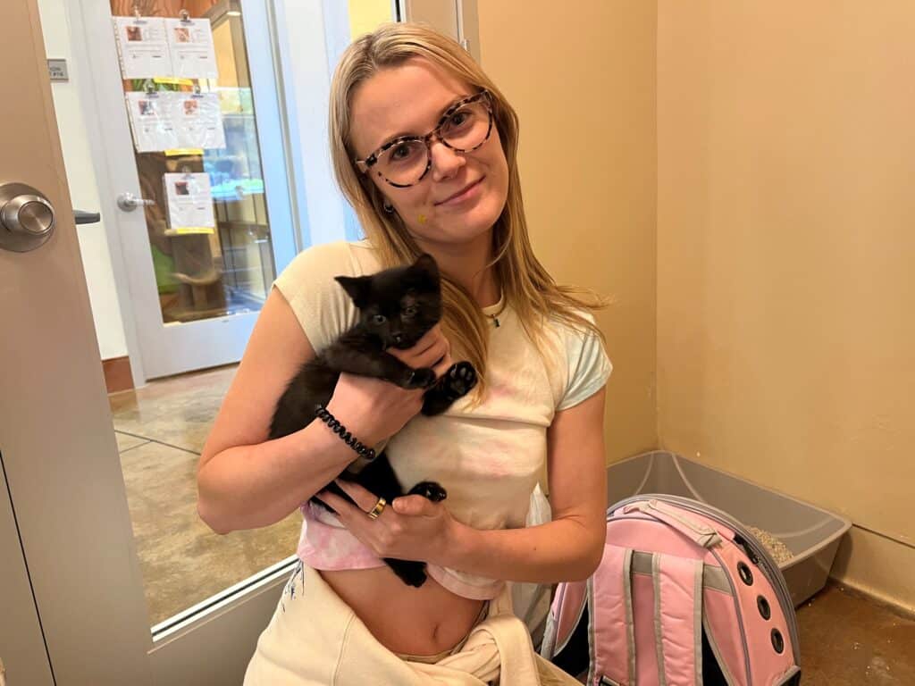 A young woman wearing glasses is smiling while holding a small black kitten in her arms. She is standing indoors next to a clear door, with a pink backpack visible on the floor nearby.