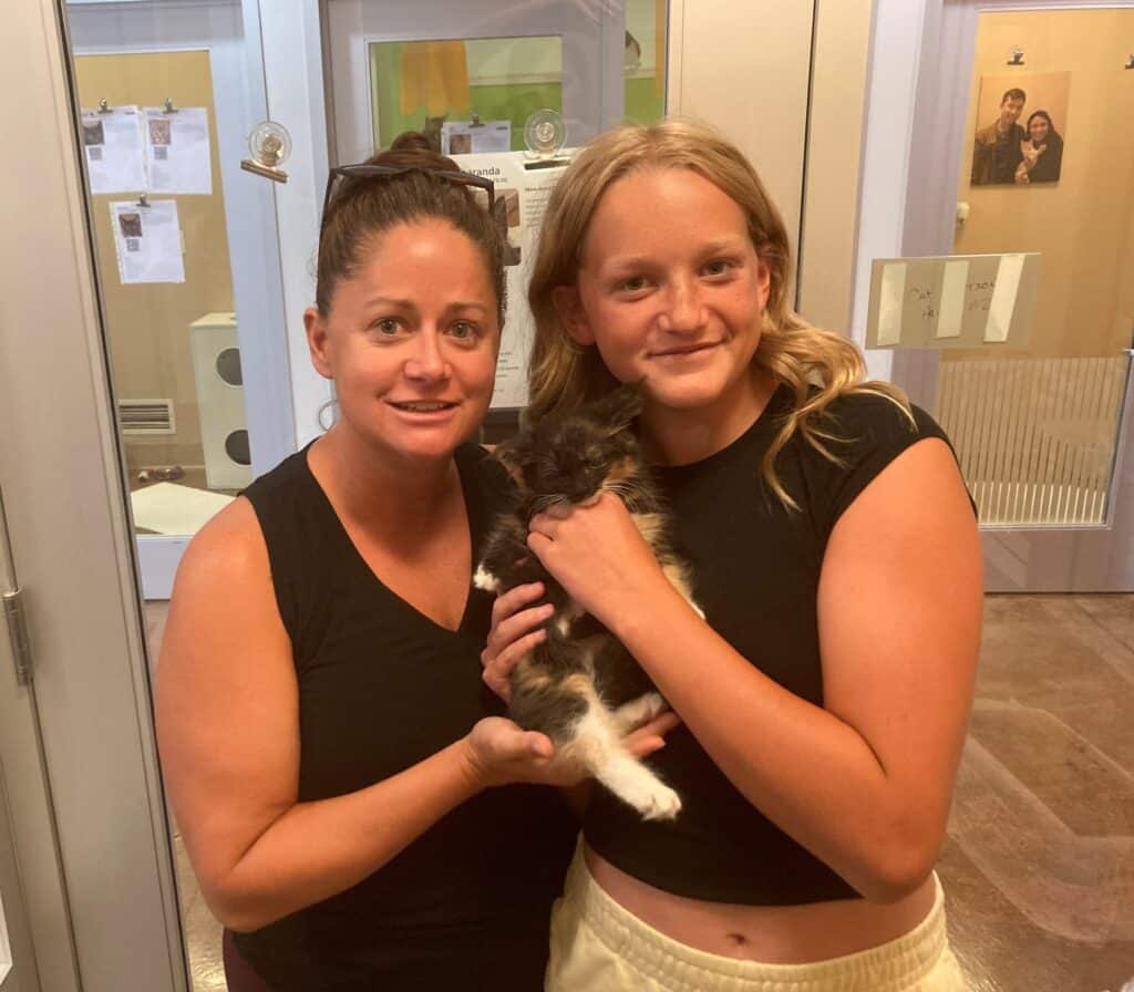 A woman and a girl are holding a small kitten together in a pet adoption center. The woman is on the left, wearing a black top, while the girl, on the right, has long hair and is wearing a black shirt. They both smile at the camera.