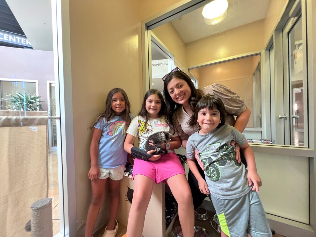 A mother and three children stand together in an adoption center. The children, two girls and a boy, are smiling. One girl holds a small pet, while the mother smiles at the camera. The environment features light-colored walls and a window.