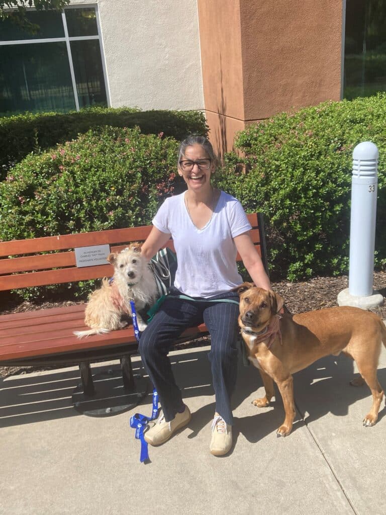 A woman sitting on a bench outside, smiling while accompanied by two dogs. The left dog is a small, light-colored terrier, and the right is a larger brown dog. The background features greenery and a building.