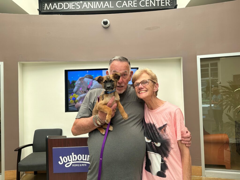 A man holding a small brown dog smiles next to a woman in a pink shirt at Maddie's Animal Care Center. A screen displaying colorful images is in the background, and a sign for Joybound Mobile Pets is visible on a nearby counter.