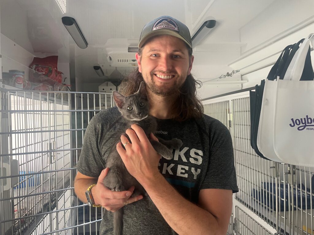 A man with long hair wears a cap and a gray t-shirt while holding a small gray kitten. They are inside an animal shelter, with cages in the background containing other cats. The setting appears bright and clean.