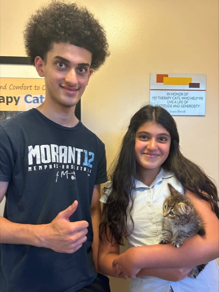 A teenage boy with curly hair gives a thumbs-up while standing next to a teenage girl holding a small kitten. The girl smiles at the camera. They are indoors near a sign referencing therapy cats.