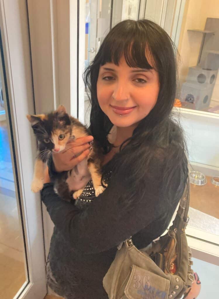 A woman holding a calico kitten while standing inside an animal shelter. The woman has long black hair and is wearing a black top. The background includes glass doors and a glimpse of other pet care areas.
