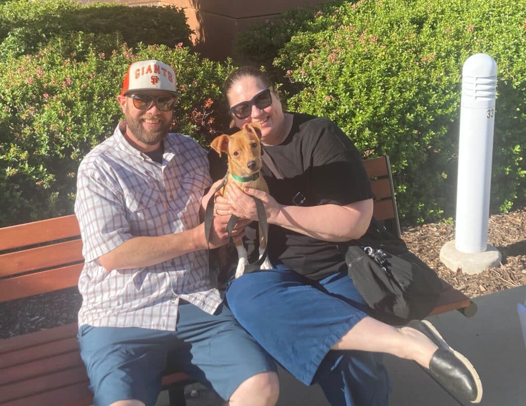 A man in a plaid shirt and a cap, and a woman wearing sunglasses, sit on a bench outdoors. They hold a small dog together, both smiling. Behind them is a backdrop of green shrubs and a white pole.