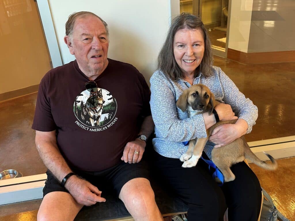 An older man wearing a black t-shirt sits beside a woman with long hair, who holds a small puppy in her arms. They are seated in a waiting area with a light-colored floor and large windows in the background.