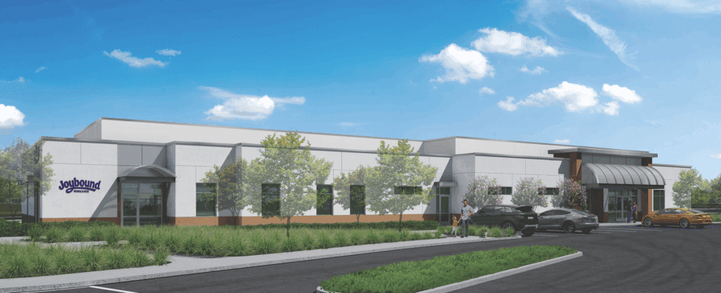 An exterior view of the Joybound People & Pets facility, showcasing a modern building with a sloped roof, surrounded by greenery and parked cars. The image represents the new dog and cat boarding and day care center, highlighting its welcoming design.