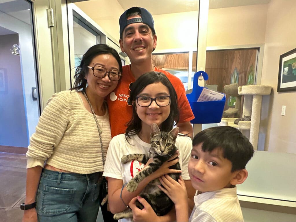 A family of four poses together in an adoption center, smiling. The young girl holds a gray striped kitten. The father stands behind them, while the mother and younger boy stand on either side, showcasing a successful adoption event. Adoptions at Joybound.