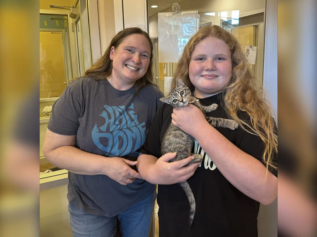 A woman and a girl smile while holding a kitten in an animal shelter setting. The woman wears a gray shirt with blue lettering, and the girl has long, wavy hair. This image represents adoptions at Joybound.