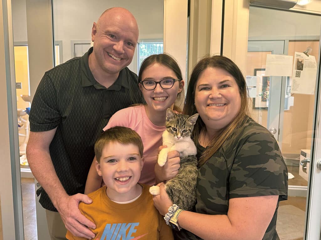 A family stands in a pet adoption center, smiling while holding a kitten. The family includes two adults and two children, with the kitten being cradled by the mother. The setting features light-colored walls and a glass door, indicating a welcoming environment. This image highlights adoptions at Joybound.