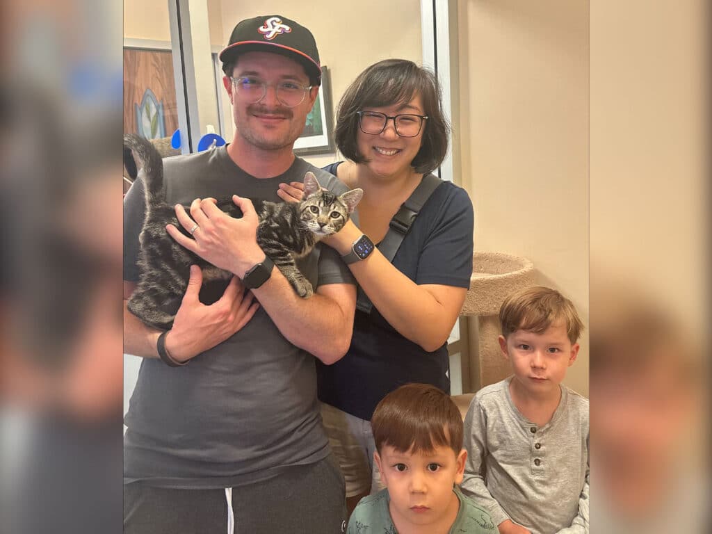 A smiling family of four poses inside a shelter. A man holds a gray tabby cat, while a woman stands beside him. Two young boys are in front of them, one looking directly at the camera. The setting appears to be a pet adoption center. Adoptions at Joybound.