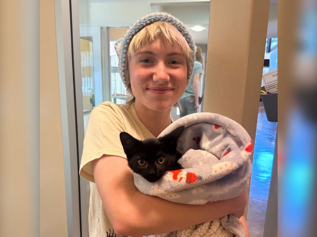 A young person wearing a light-colored shirt and a knit hat smiles while holding a black kitten wrapped in a gray blanket with red and white patterns. The setting appears to be indoors, likely a shelter or adoption event related to Adoptions at Joybound.